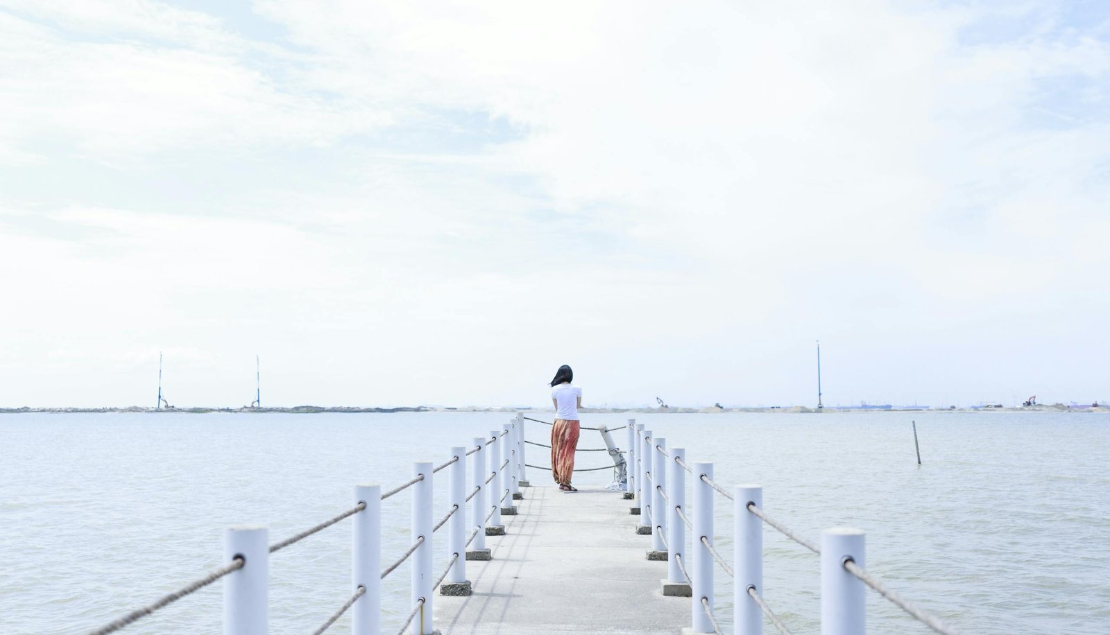 A woman stands quietly on a serene pier overlooking the calm ocean under a cloudy sky.